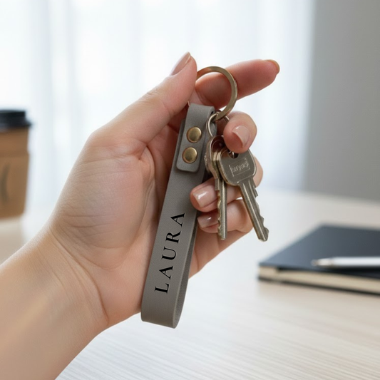 Rankoje laikomas juodas odinis rakto pakabukas su metaliniu žiedu ir raktų ryšuliu – minimalistinis aksesuaras el. parduotuvei