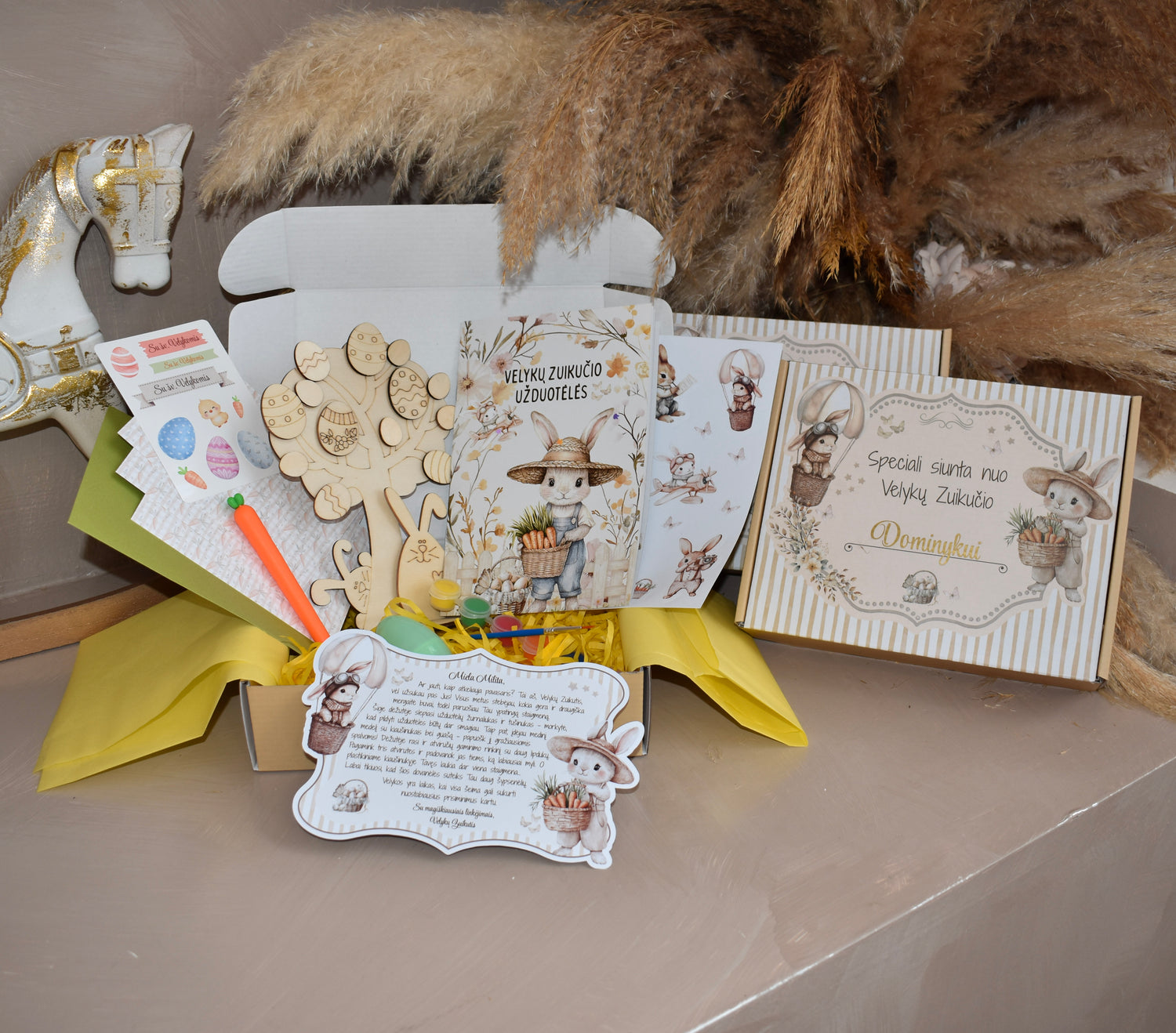 Atvira Velykų zuikučio dovanų dėžutė vaikams ant šviesaus pagrindo, apsupta tulpių ir margučių. Dėžutės viduje matomas užduotėlių žurnalas su kiškučiu, tušinukas-morkytė, medinis kiaušinių medelis, guašas, atvirutės ir lipdukai.